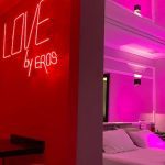 Quarto temático com iluminação neon vermelha, letreiro 'LOVE by EROS' e cama com roupa de cama branca.