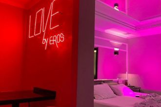 Quarto temático com iluminação neon vermelha, letreiro 'LOVE by EROS' e cama com roupa de cama branca.