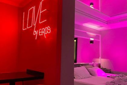 Quarto temático com iluminação neon vermelha, letreiro 'LOVE by EROS' e cama com roupa de cama branca.