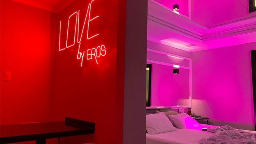 Quarto temático com iluminação neon vermelha, letreiro 'LOVE by EROS' e cama com roupa de cama branca.