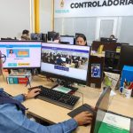 Servidores trabalhando em computadores em escritório, com destaque para telas mostrando o site da prefeitura de Manaus e um livro sobre concursos públicos.