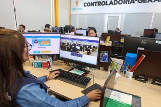 Servidores trabalhando em computadores em escritório, com destaque para telas mostrando o site da prefeitura de Manaus e um livro sobre concursos públicos.