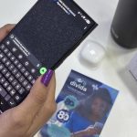 Uma pessoa usando um celular com teclado na tela, unhas roxas e outros objetos na mesa.