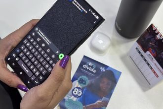 Uma pessoa usando um celular com teclado na tela, unhas roxas e outros objetos na mesa.