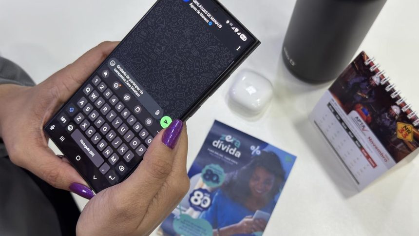 Uma pessoa usando um celular com teclado na tela, unhas roxas e outros objetos na mesa.