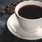 Xícara branca com café fumegante e grãos de café em fundo escuro.