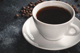 Xícara branca com café fumegante e grãos de café em fundo escuro.