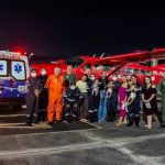 Grupo posa em frente a avião vermelho e ambulância. Mulheres com bebês e equipes de resgate.