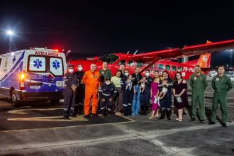 Grupo posa em frente a avião vermelho e ambulância. Mulheres com bebês e equipes de resgate.