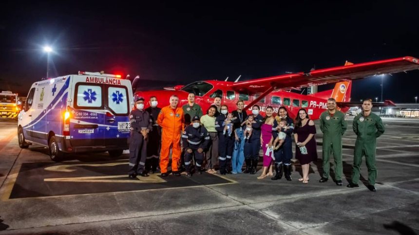 Grupo posa em frente a avião vermelho e ambulância. Mulheres com bebês e equipes de resgate.