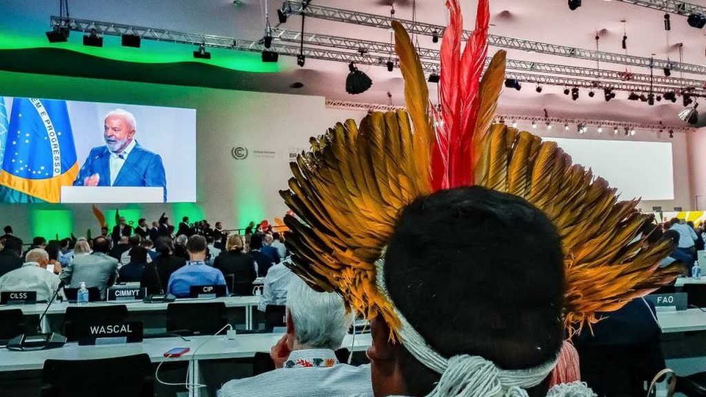 Indígena com cocar em evento com Lula falando em painel e bandeira do Brasil