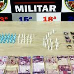 Apreensão de drogas e dinheiro pela Polícia Militar. Itens apreendidos incluem pinos, embalagens e cédulas.