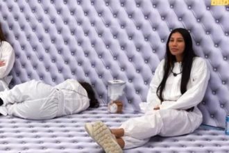 Participantes no interior da casa do Big Brother Brasil, vestindo macacões brancos, no quarto branco