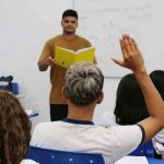 Professor em sala de aula com alunos, um deles com a mão levantada para responder
