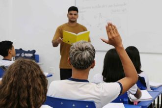 Professor em sala de aula com alunos, um deles com a mão levantada para responder