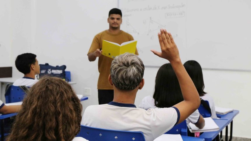 Professor em sala de aula com alunos, um deles com a mão levantada para responder
