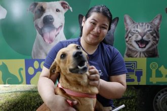 Mulher sorrindo segura um cachorro de pêlo marrom com uma coleira vermelha, com rostos de animais ao fundo.