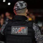 Policial militar de costas com uniforme camuflado e a palavra ROCAM no colete.