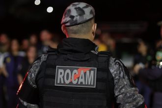 Policial militar de costas com uniforme camuflado e a palavra ROCAM no colete.