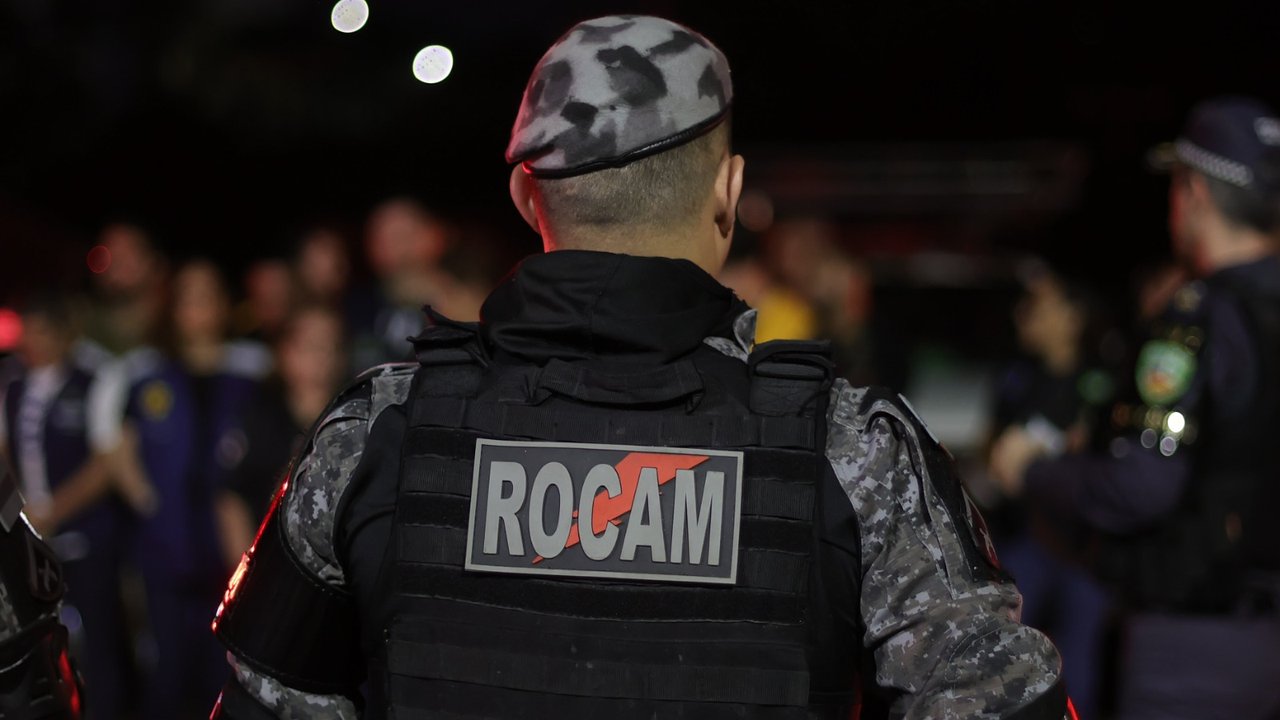 Policial militar de costas com uniforme camuflado e a palavra ROCAM no colete.