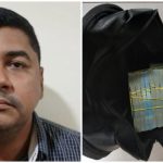 Policial Militar do Amazonas ao lado de mala preta aberta com maços de dinheiro amarrados.