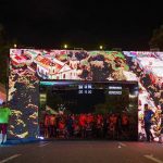 Largada da Maratona Internacional de Manaus com tela digital mostrando imagens, pessoas e relógios.