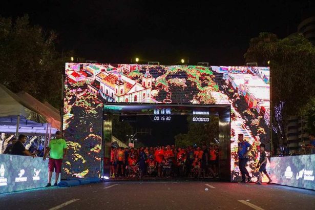 Largada da Maratona Internacional de Manaus com tela digital mostrando imagens, pessoas e relógios.