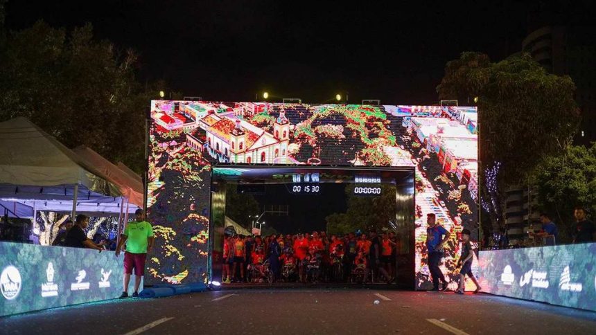 Largada da Maratona Internacional de Manaus com tela digital mostrando imagens, pessoas e relógios.