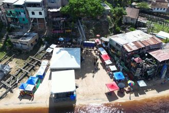 Vista aérea da praia da Ponta Branca com barracas, prédios coloridos e barcos.
