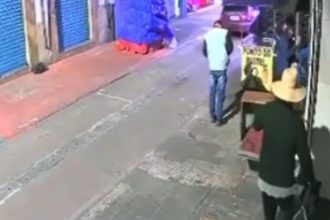 Cena de rua com pessoas, calçada e fachada de loja. Um homem caminha na calçada.