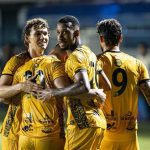 Jogadores do Amazonas FC celebrando um gol, abraçados e vestindo uniformes amarelos.