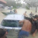 Pessoas ajudando em um carro em meio a uma enchente, com barco próximo e chuva forte.