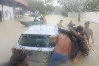 Pessoas ajudando em um carro em meio a uma enchente, com barco próximo e chuva forte.