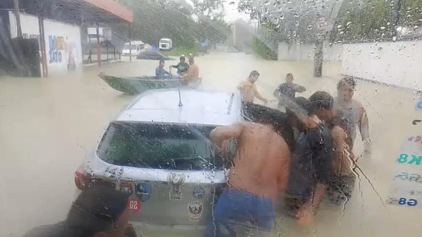 Pessoas ajudando em um carro em meio a uma enchente, com barco próximo e chuva forte.