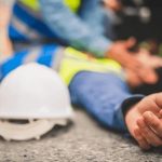 Acidente de trabalho: trabalhador ferido no chão, com capacete próximo e outros funcionários prestando socorro.