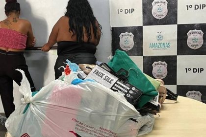Duas mulheres em uma sala com evidências apreendidas, incluindo roupas e sacolas plásticas.
