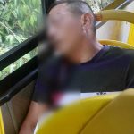 Homem sentado em um ônibus amarelo, com rosto desfocado, com marcas de tiros, ao lado de janelas com vegetação.