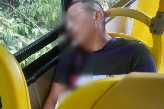 Homem sentado em um ônibus amarelo, com rosto desfocado, com marcas de tiros, ao lado de janelas com vegetação.