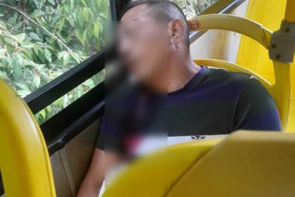 Homem sentado em um ônibus amarelo, com rosto desfocado, com marcas de tiros, ao lado de janelas com vegetação.