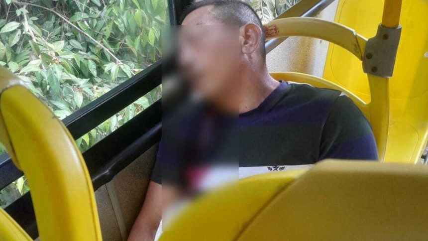 Homem sentado em um ônibus amarelo, com rosto desfocado, com marcas de tiros, ao lado de janelas com vegetação.