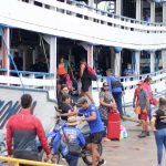 Pessoas embarcando e desembarcando de um barco em um píer, com um céu nublado ao fundo.